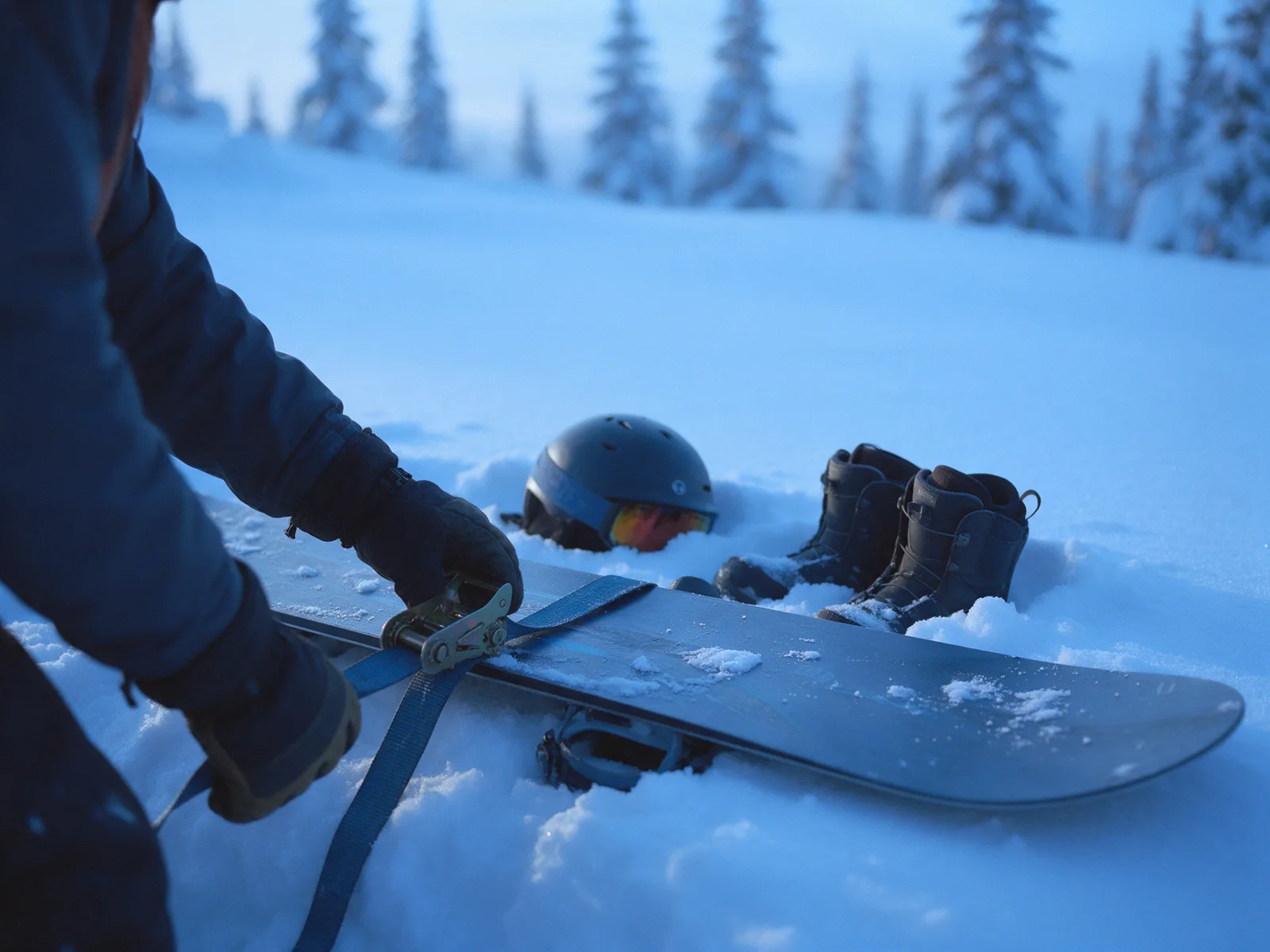 Step 2 &ndash; Hands adjusting a snowboard binding in the snow, with a helmet and boots alongside