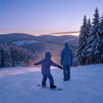 Kind auf einem kurzen Snowboard gleitet mit ausgebreiteten Armen einen flachen Hang hinunter, daneben ein erwachsener Snowboardlehrer